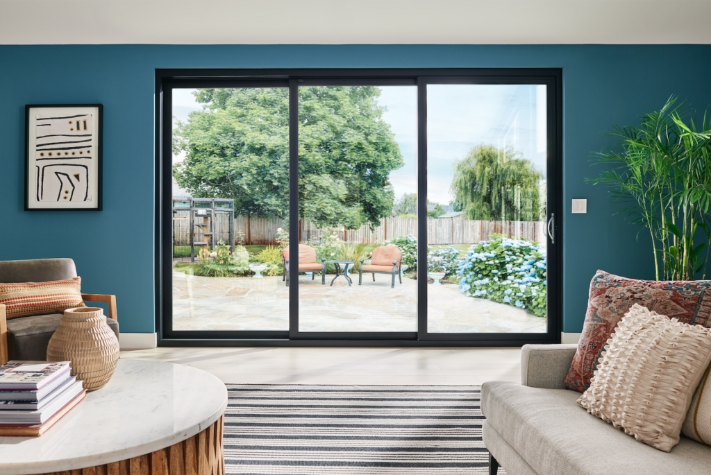 wide shot of a three-paned sliding glass door inside a room with a blue wall looking out into a sitting area with green trees on the outside