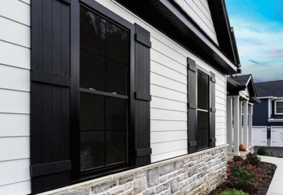 Close-up view of the side of a modern residential house featuring a combination of vertical white siding and stone veneer at the base. The house has large windows with dark frames and shutters, enhancing the stylish contrast against the white siding