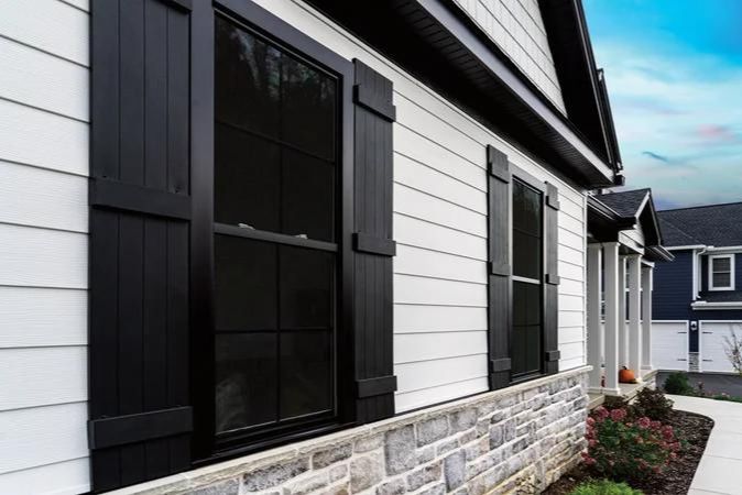 Close-up view of the side of a modern residential house featuring a combination of vertical white siding and stone veneer at the base. The house has large windows with dark frames and shutters, enhancing the stylish contrast against the white siding