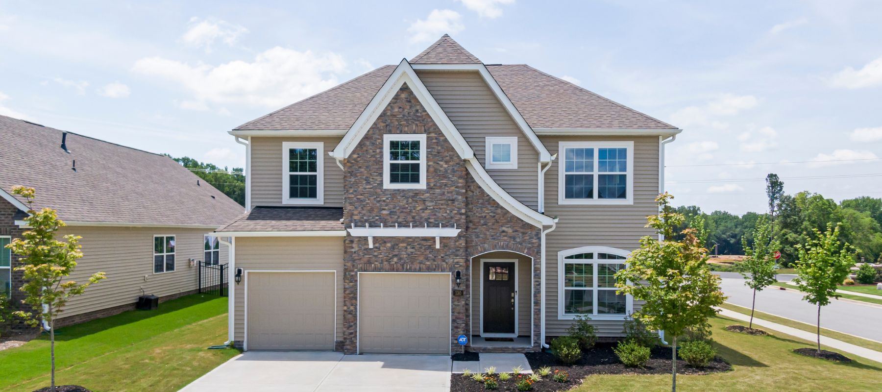 A large two-story suburban house features a mix of tan siding and stone veneer, with two garage doors and a prominent front entrance, under a partly cloudy sky