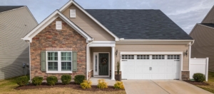 A cozy single-story house with tan siding and stone veneer features a white garage door, green shutters, and a dark front door with a wreath